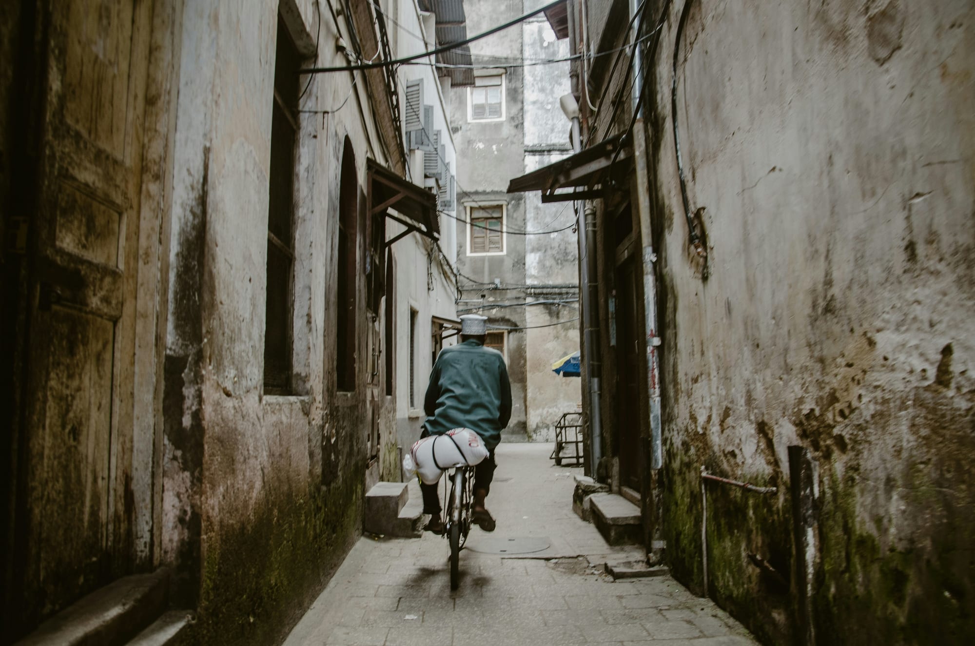 A cyclist riding down a narrow lane between weathered stone buildings