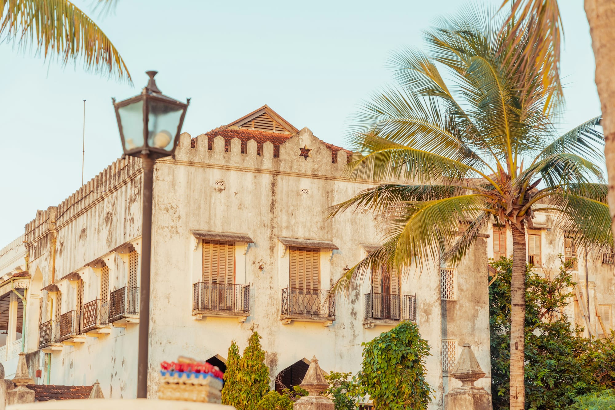 A weathered stone building with carved balconies behind palm trees