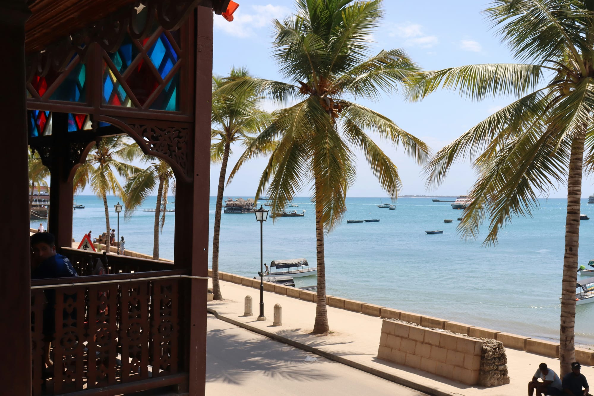 A carved wooden balcony looking out to palms, boats and open sea